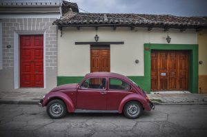 A vintage red volkswagen beetle parked on a cobblestone street in front of two colorful buildings with red and green doors.