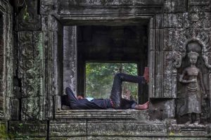 A person lies on a stone ledge of an ancient window frame amidst ruins in Cambodia, with elaborate carvings and a statue visible on the surrounding walls.