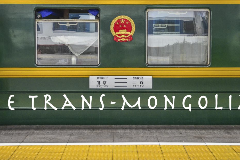 A green train, featured on a Travel Blog, with "the trans-mongolian" text on its side is stopped at a platform, with national emblem decals indicating its international route between Beijing and U